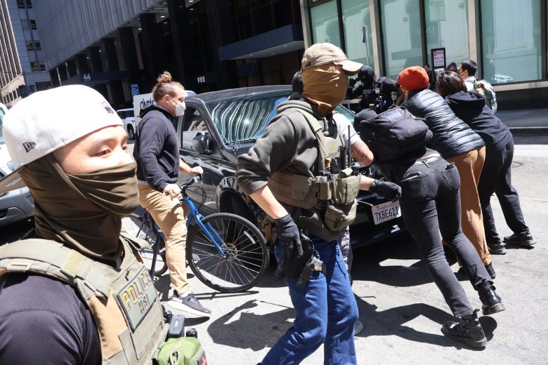 People in tactical gear detain several individuals against a black SUV on a city street while others observe; a bicycle is visible in the background.