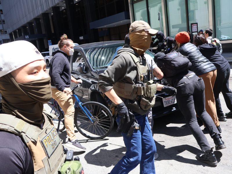 People in tactical gear detain several individuals against a black SUV on a city street while others observe; a bicycle is visible in the background.