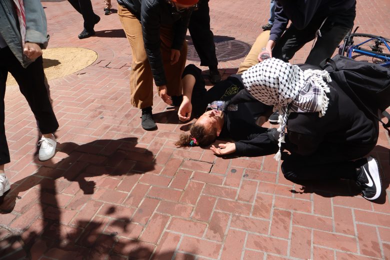Several people assist a person lying on a brick sidewalk; a bicycle is nearby and bystanders are present.