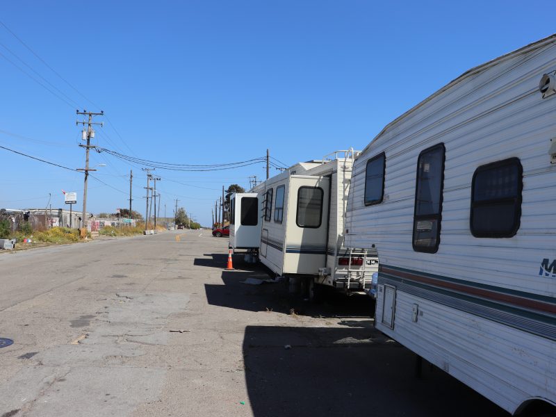Several RVs are parked along the side of an empty urban street in designated parking spots under a clear blue sky. Power lines and buildings run alongside the road.