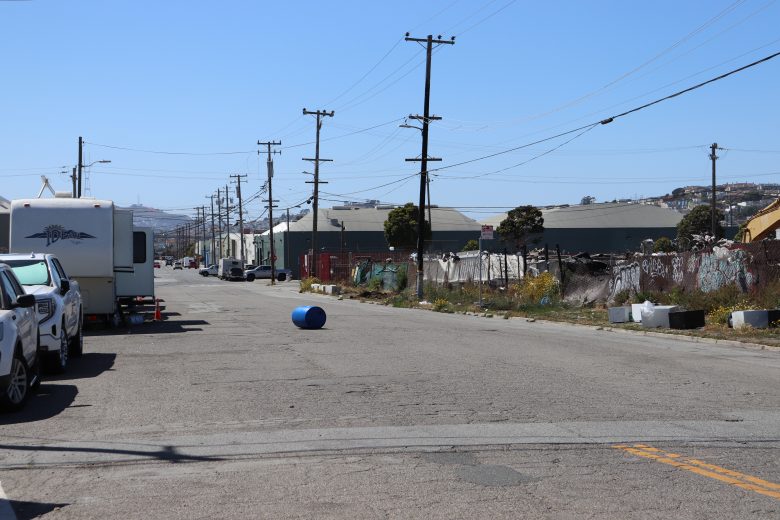 A blue barrel lies in the middle of an empty industrial street lined with parked vehicles, utility poles, and buildings on a clear day.