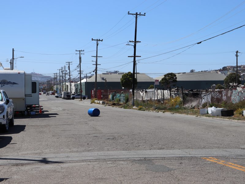 A blue barrel lies in the middle of an empty industrial street lined with parked vehicles, utility poles, and buildings on a clear day.
