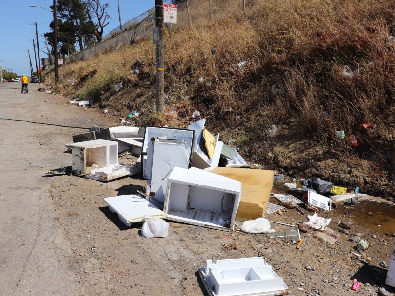 Discarded household appliances and trash litter a roadside next to a grassy embankment; a person in a yellow vest is visible in the distance.
