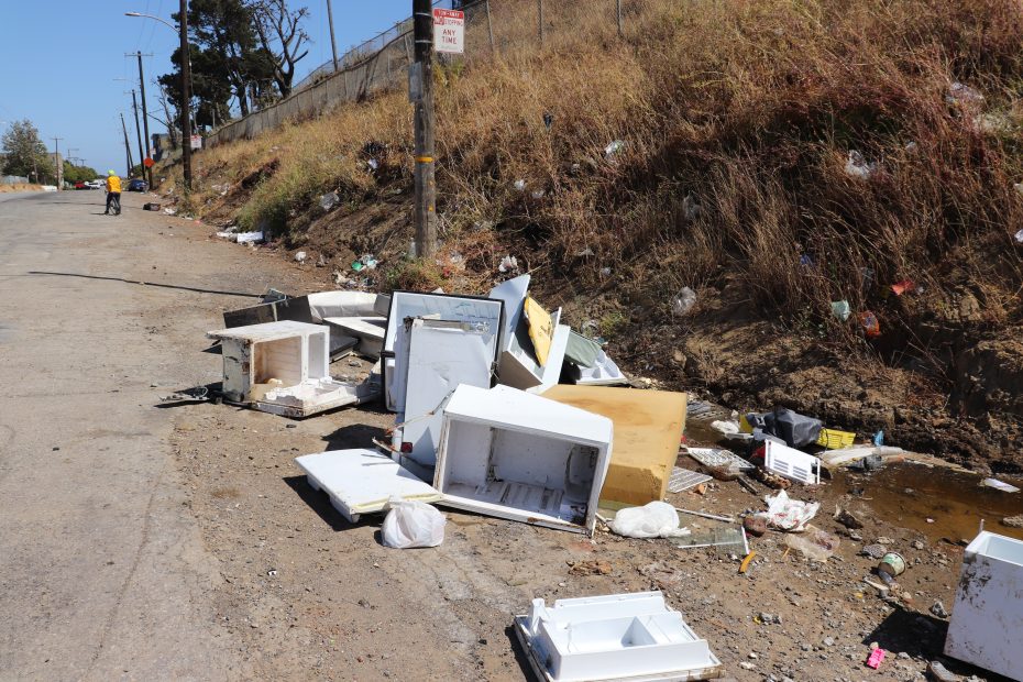 Discarded household appliances and trash litter a roadside next to a grassy embankment; a person in a yellow vest is visible in the distance.