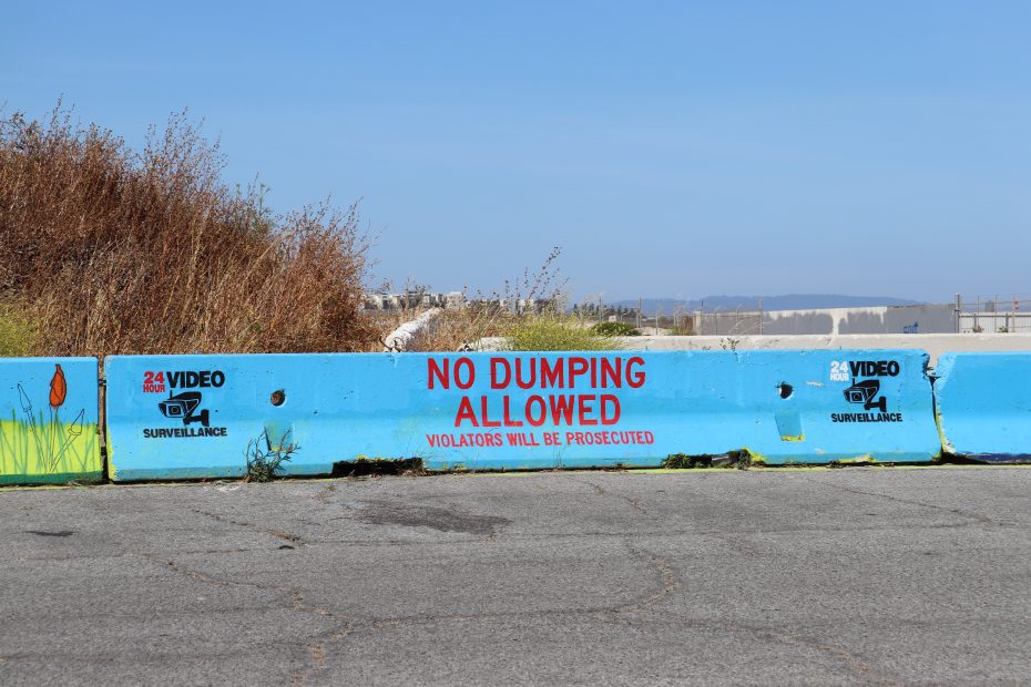 Concrete barrier with "No Dumping Allowed" and "24h Video Surveillance" signs, placed on an asphalt surface with weeds and brush in the background under a clear sky.