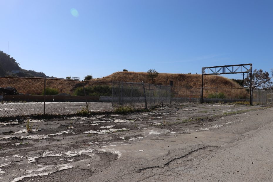 Abandoned fenced lot with cracked pavement, overgrown weeds, and a grassy hill in the background under a clear blue sky.