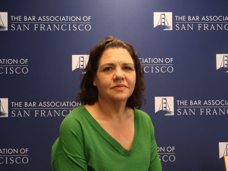 A woman with medium-length brown hair in a green top sits in front of a backdrop with "The Bar Association of San Francisco" logos.