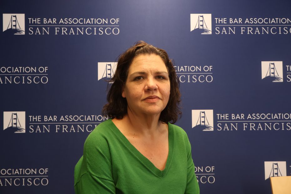 A woman with medium-length brown hair in a green top sits in front of a backdrop with "The Bar Association of San Francisco" logos.