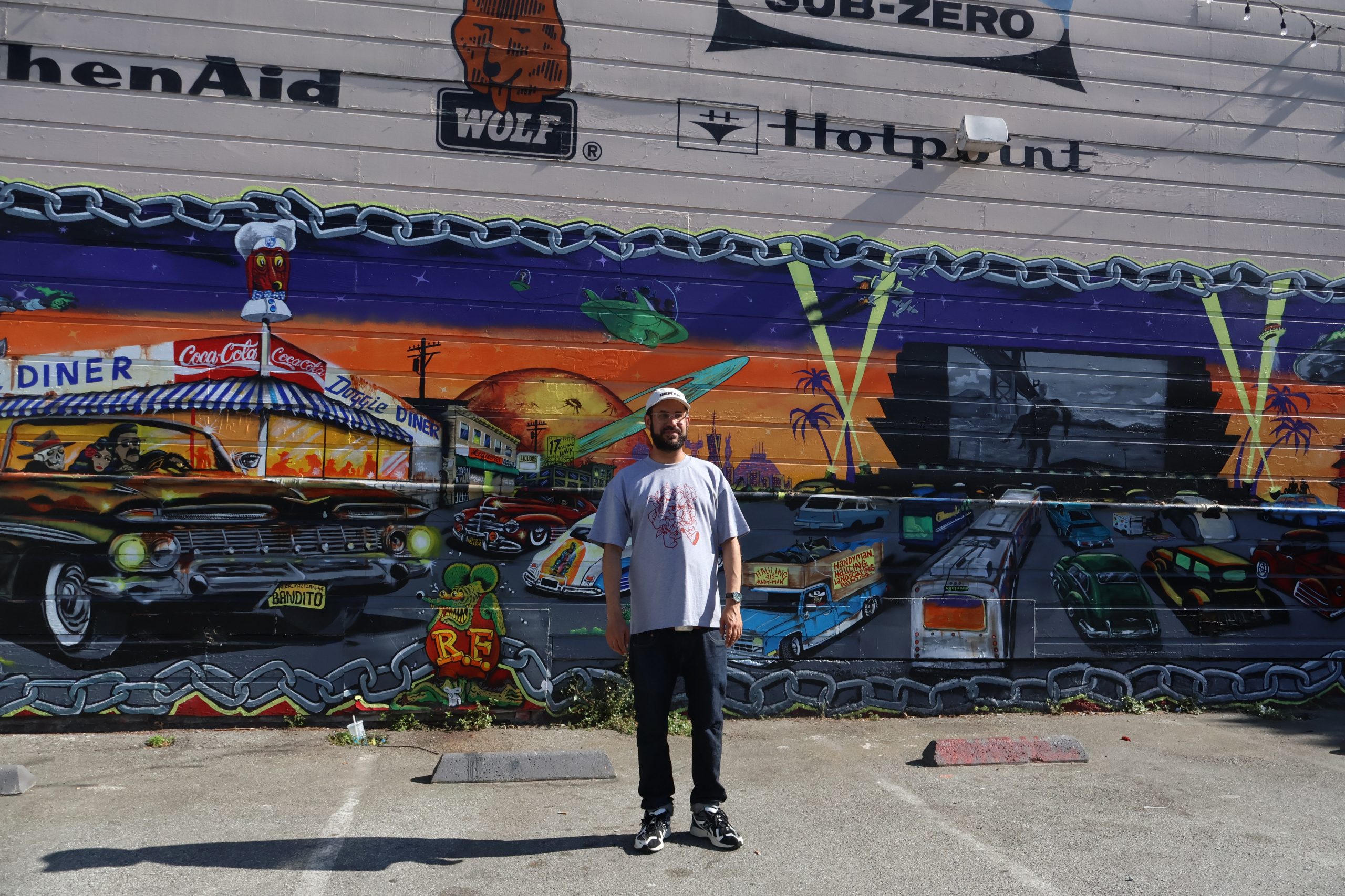 A man stands in front of a colorful mural depicting classic cars, a diner, and various retro scenes, painted on the side of a building in a parking lot.