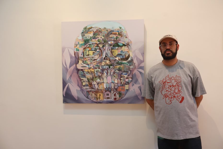 A man in a gray t-shirt and cap stands next to a painting of a skull filled with images of houses and buildings, displayed on a white wall.