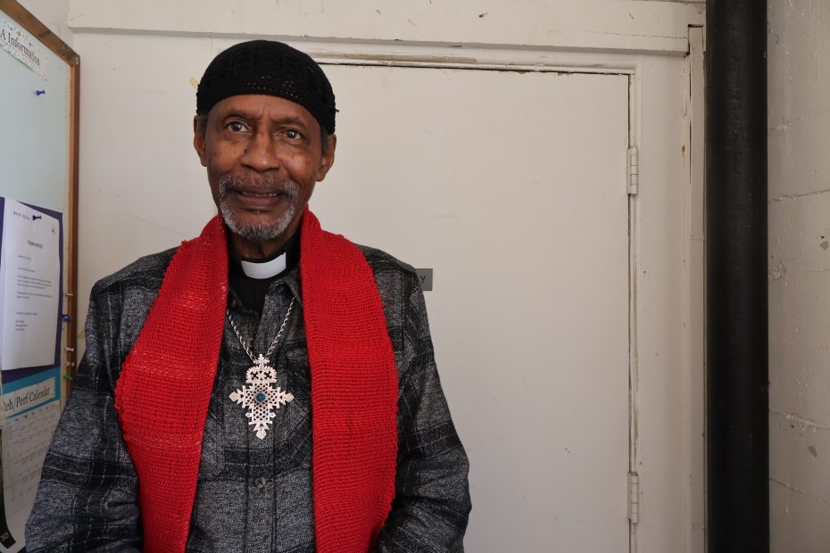 An older man wearing a red scarf, black cap, and large cross necklace stands indoors near a closed white door and a bulletin board.
