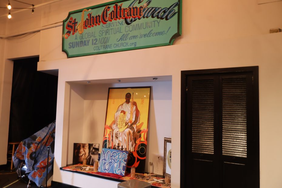 A portrait of a seated man with a saxophone is displayed beneath a sign reading "St. John Coltrane Church" in a room with various pictures and objects.