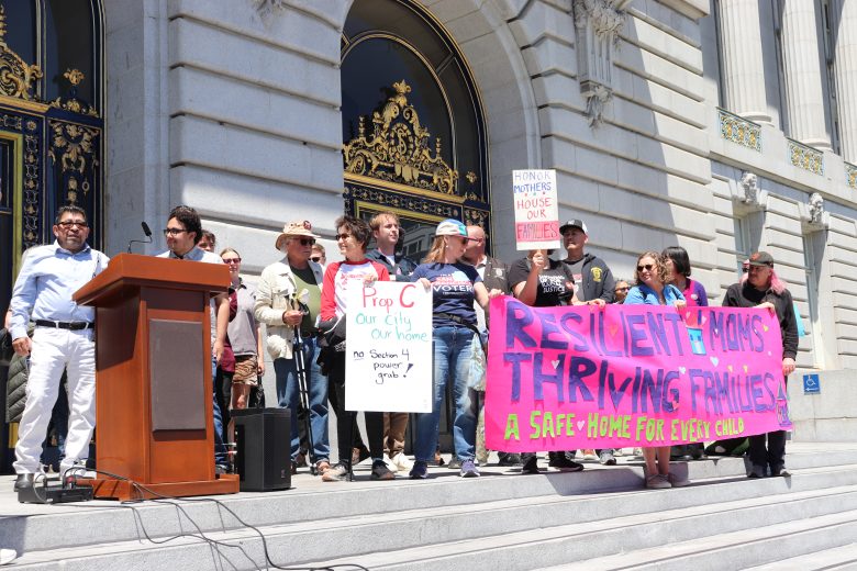 A group of people stand on city hall steps holding signs and a pink banner that reads, “RESILIENT MOMS. THRIVING FAMILIES. A SAFE HOME FOR EVERY CHILD.”.
