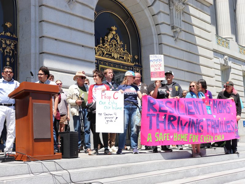 A group of people stand on city hall steps holding signs and a pink banner that reads, “RESILIENT MOMS. THRIVING FAMILIES. A SAFE HOME FOR EVERY CHILD.”.