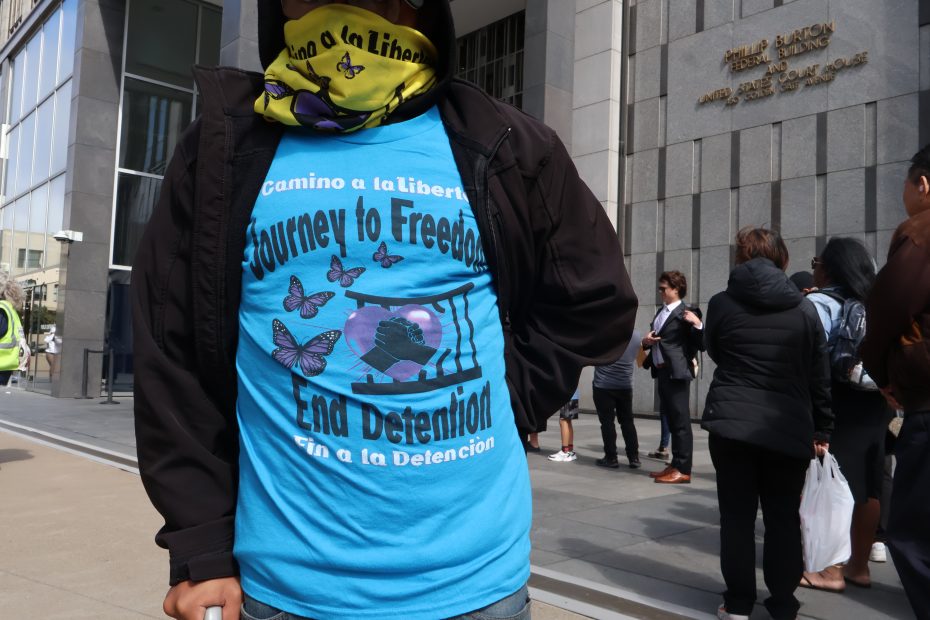 Person wearing a blue shirt with text "Journey to Freedom, End Detention" and butterfly graphics, standing outside a building with other people in the background.