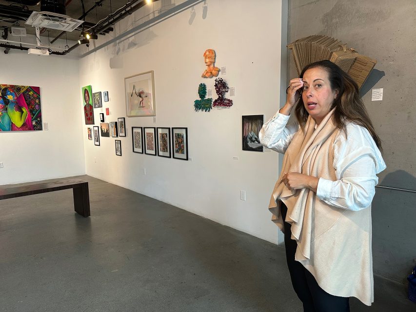 A woman stands in an art gallery, gesturing near her head, with various colorful artworks displayed on the white wall behind her.