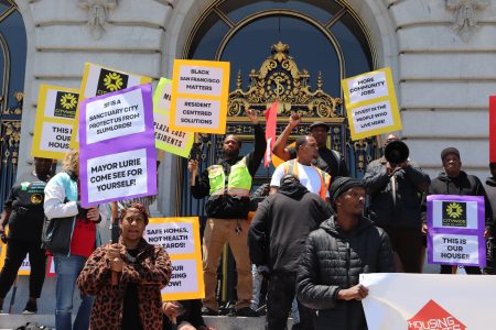 A group of people stand on building steps in San Francisco, holding signs advocating for housing rights and community issues.