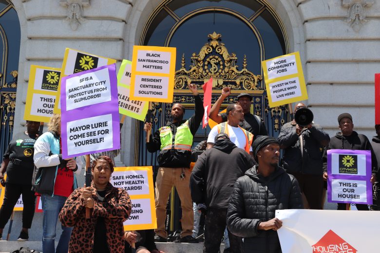 A group of people stand on building steps in San Francisco, holding signs advocating for housing rights and community issues.