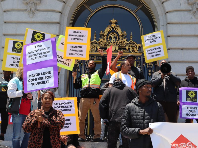 A group of people stand on building steps in San Francisco, holding signs advocating for housing rights and community issues.