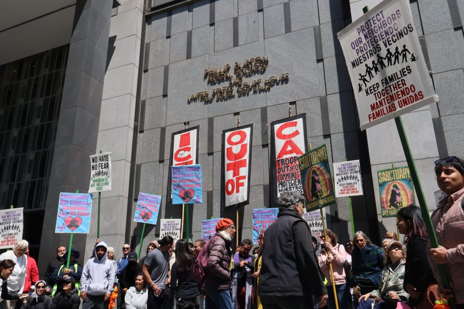 A group of people hold protest signs reading "ICE OUT OF CA" and other messages in front of a gray government building labeled "United States Court House.