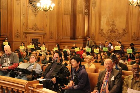 People seated in a wood-paneled room, some in wheelchairs, while others stand holding yellow signs in the back. Chandelier lighting is visible above.