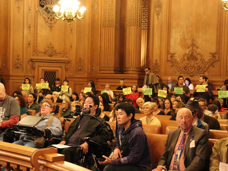 People seated in a wood-paneled room, some in wheelchairs, while others stand holding yellow signs in the back. Chandelier lighting is visible above.