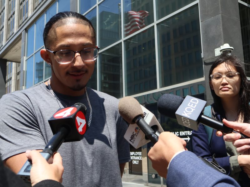 A man speaks to reporters holding microphones outside a building with glass windows; an American flag is visible in the reflection.