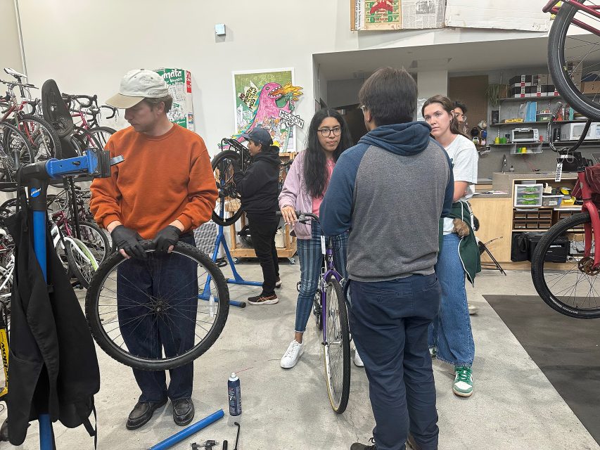A group of people work together to repair bicycles in a workshop, surrounded by tools, bike parts, and bikes mounted on stands.