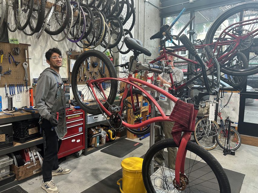 A mechanic stands in a bike repair shop next to a red bicycle mounted on a stand, surrounded by various tools and bikes hanging on the wall and ceiling.