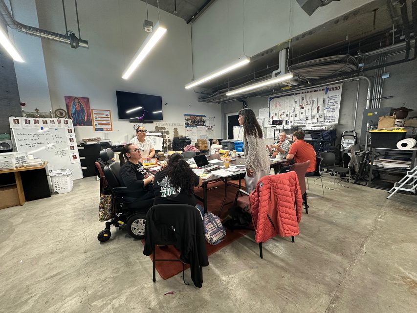 A group of people sit and work at a rectangular table in an industrial-style room with papers, laptops, and jackets on the table. A whiteboard and posters are visible in the background.