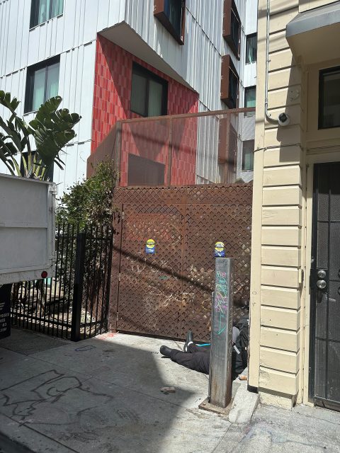 A person lies on a sidewalk near a brown metal gate outside an urban building; a white truck is parked nearby, and graffiti is visible on a pole and ground.