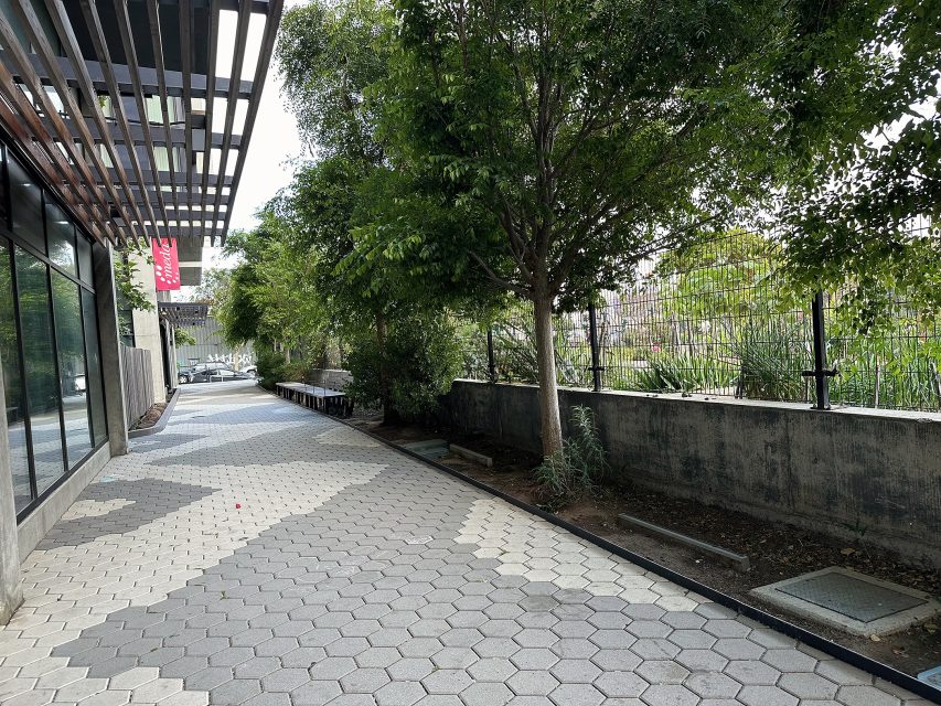 A paved walkway lined with trees runs alongside Mission, a building with glass windows and a metal awning. A concrete wall and garden beds are on the right.