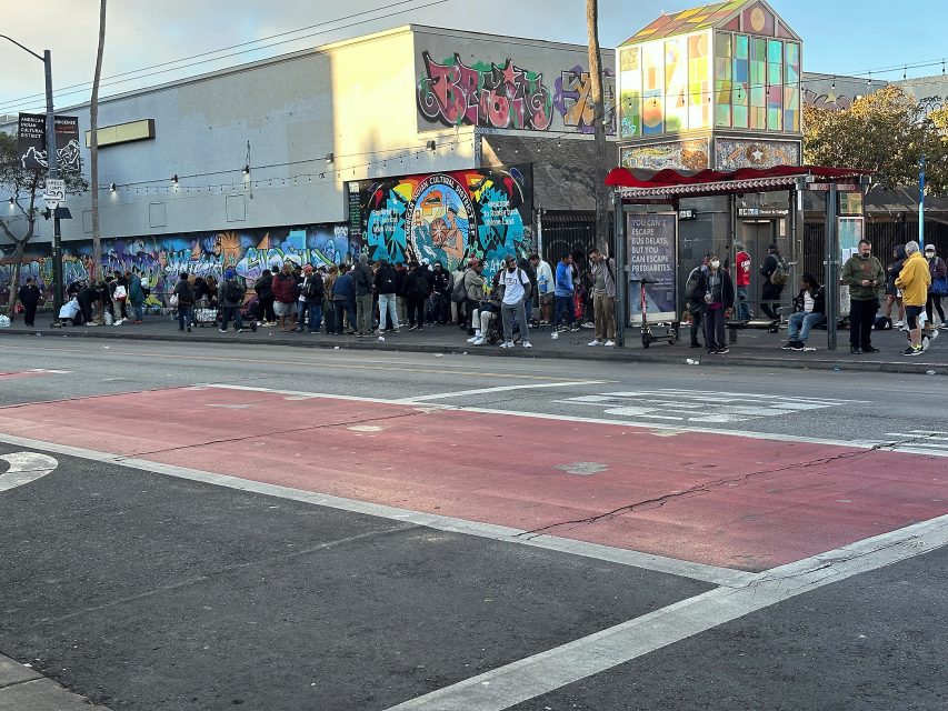 A long line of people waits outside a building with colorful graffiti murals and a glass-paneled structure at a city street intersection.