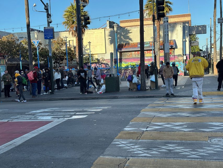 A group of people stand in line on a city sidewalk near a mural, while a person in a yellow jacket walks across a crosswalk in the foreground.