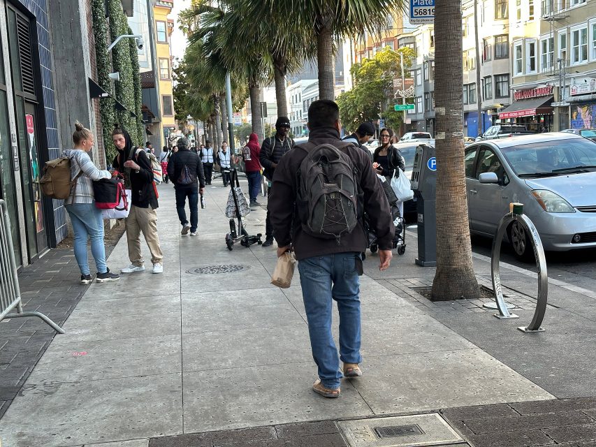 A man with a backpack walks on a city sidewalk; groups of people stand nearby, scooters are parked, and cars line the street on a sunny day.
