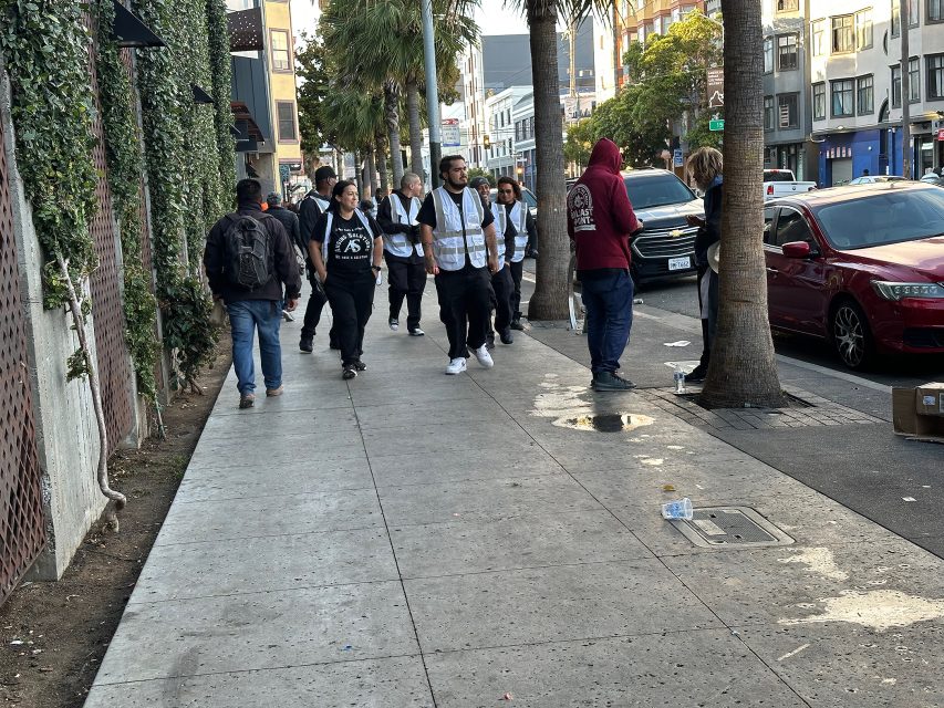 A group of people, some wearing reflective vests, walk on a city sidewalk lined with palm trees and parked cars.