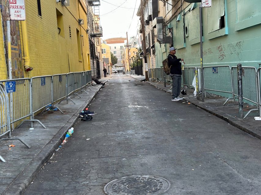A man stands in an empty alleyway lined with metal barricades and trash. Buildings on both sides have graffiti and broken windows.