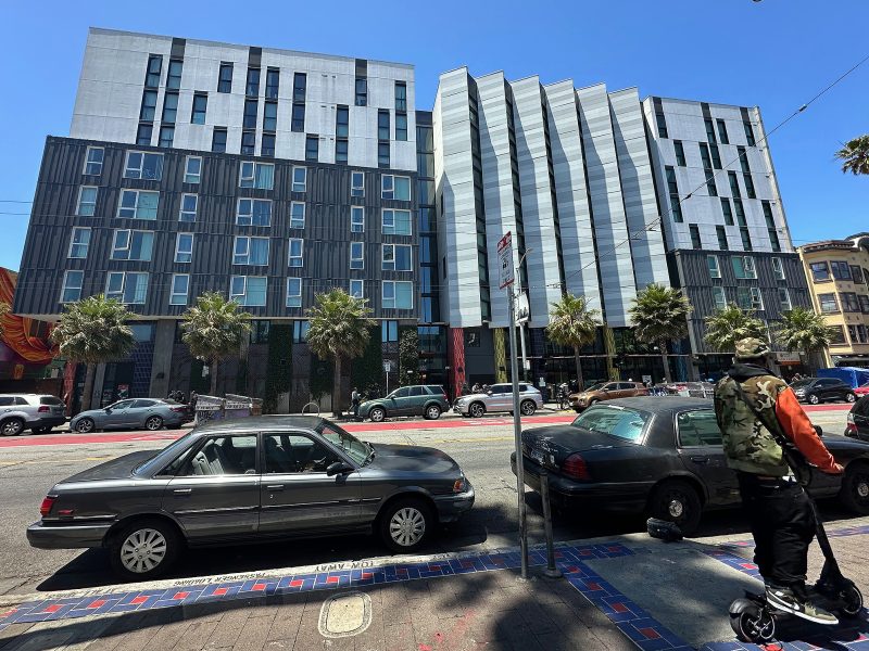 A street view of a modern multi-story building with parked cars, palm trees, and a person riding a scooter in the foreground.