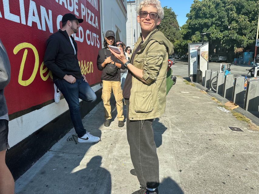A group of people stand and interact on a sunny sidewalk near a building with a red and white sign, while rental bikes are visible in the background.