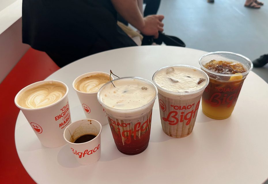 Five assorted coffee drinks, both hot and cold, are arranged on a round white table. Some people are seated in the background.