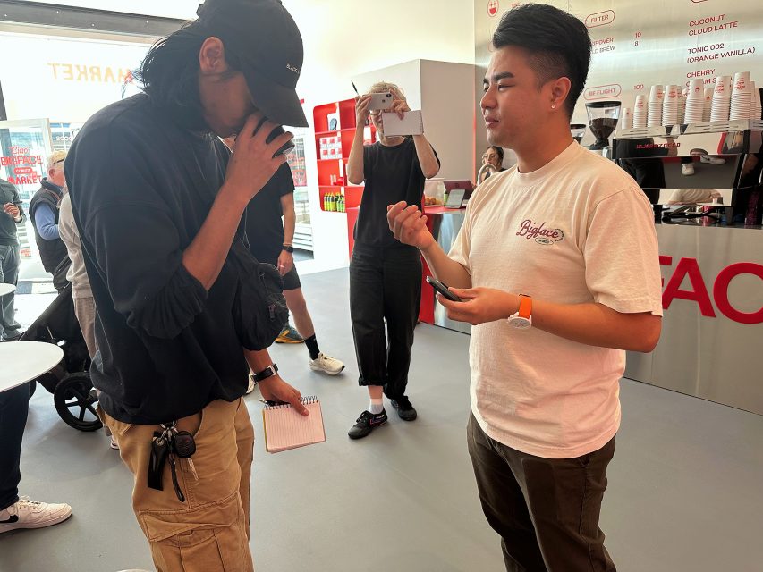 A man in a black outfit smells a cup while another man in a white t-shirt speaks to him; people and coffee equipment are visible in the background.
