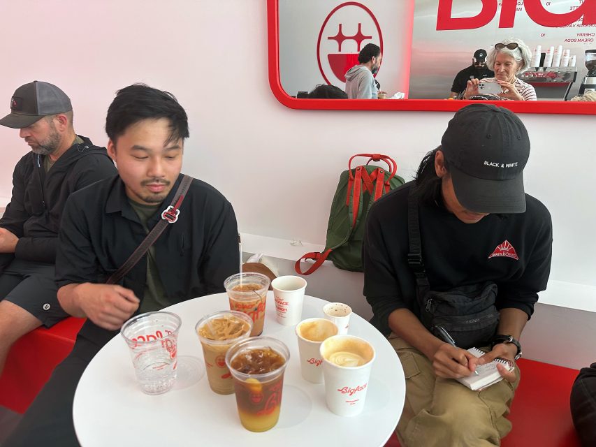 Two people sit next to a small table with multiple cups of coffee and iced drinks at a cafe; a mirror behind them shows others and the cafe’s interior.