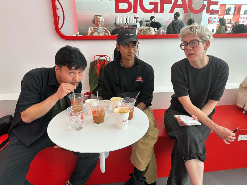 Three people sit at a round table with drinks in a modern café. One drinks with a straw, another looks ahead, and the third holds a notepad. The word "BIGFACE" is visible in the background.