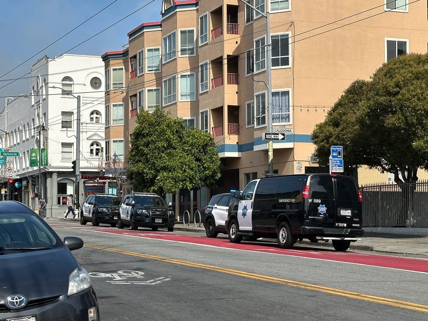 Three police vehicles are parked along a city street in front of an apartment building, with a red bus lane and bike lane markings visible.