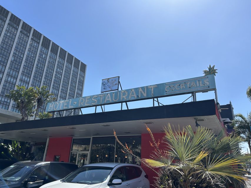 A low-rise building with a sign reading "HOTEL - RESTAURANT COCKTAILS" under a clear blue sky, with parked cars and palm trees in front.