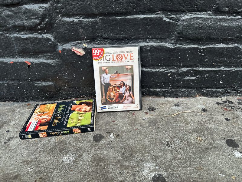 Two DVD cases, one upright and one flat, are on a sidewalk against a black brick wall. The upright case is for "GLOW: The Complete Second Season.