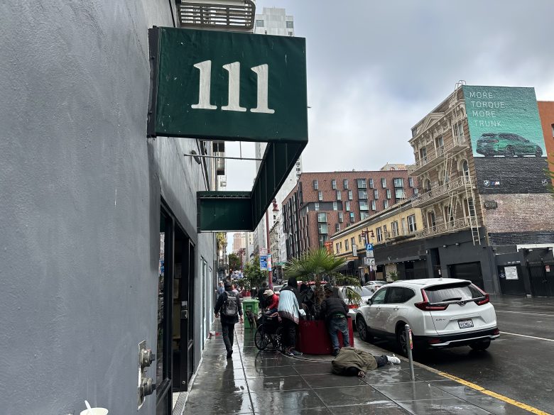 A green awning with the number 111 hangs over a wet city sidewalk where people and parked cars line the street on a cloudy day.