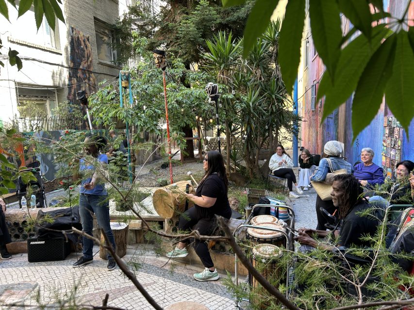 A group of people gathers outdoors among plants; some play drums and percussion instruments while others watch or sit nearby on benches.
