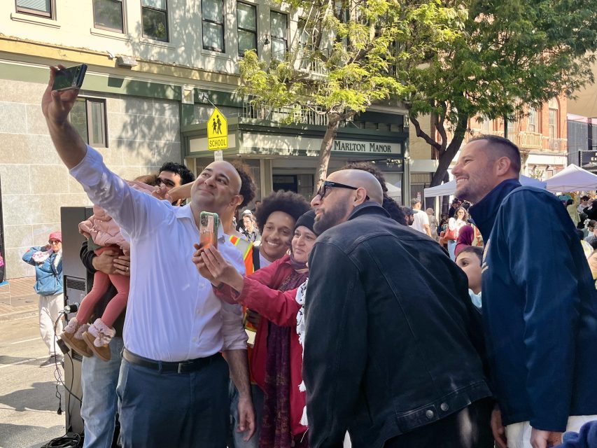 A group of people, including adults and a child, stand on a city street taking a group selfie in front of a building and a street sign.