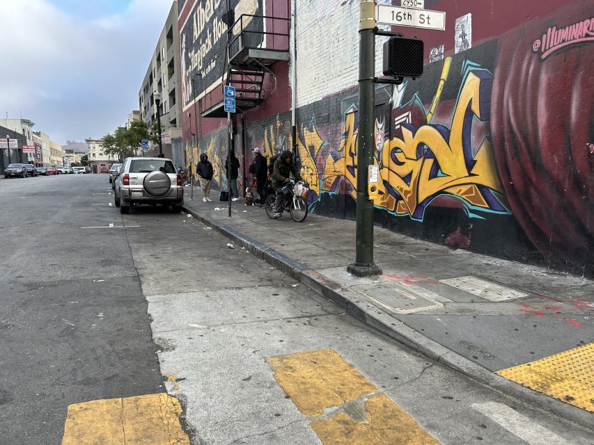 People stand near a wall with graffiti art by a street corner, next to parked cars and a traffic signal at the intersection of 16th Street.